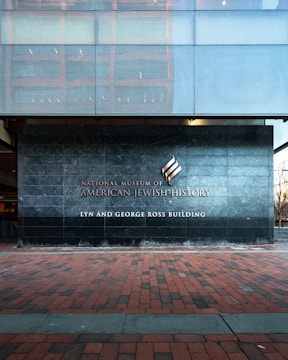 A modern building facade with a large stone wall displaying the name 'National Museum of American Jewish History, Lyn and George Ross Building.' The ground is covered with red and gray brick tiles, and the upper section features reflective glass panels.