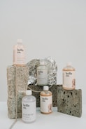 A collection of hair treatment products from 'hella co.' are arranged artfully on a surface. The bottles are labeled with different flavors like Grapefruit, Blueberry, and Kiwifruit, each with a minimalist design. They are set against a background of rustic stones and foil, with a string of beads adding a decorative touch.