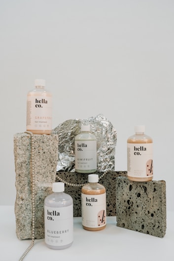 A collection of hair treatment products from 'hella co.' are arranged artfully on a surface. The bottles are labeled with different flavors like Grapefruit, Blueberry, and Kiwifruit, each with a minimalist design. They are set against a background of rustic stones and foil, with a string of beads adding a decorative touch.