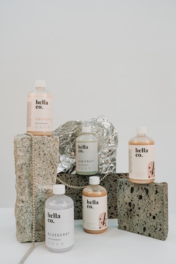 A collection of hair treatment products from 'hella co.' are arranged artfully on a surface. The bottles are labeled with different flavors like Grapefruit, Blueberry, and Kiwifruit, each with a minimalist design. They are set against a background of rustic stones and foil, with a string of beads adding a decorative touch.