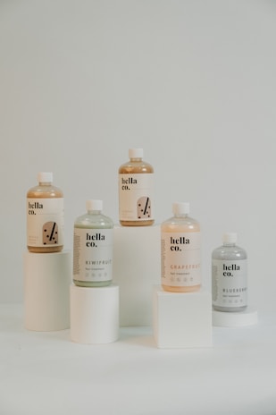 Five bottles of hair treatment products with labels displaying different fruit flavors are arranged on cylindrical white pedestals. The bottles are labeled 'hella co.' and include flavors such as grapefruit, kiwifruit, and blueberry.