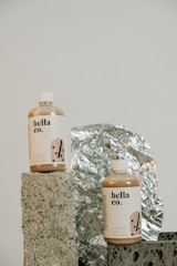 Two bottles labeled 'hella co.' with designs indicating milk tea products, one for shower cream and the other for hair therapy, are placed on textured stone-like blocks. A reflective, crinkled foil serves as the backdrop, adding a metallic element to the composition.