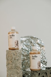 Two bottles labeled 'hella co.' with designs indicating milk tea products, one for shower cream and the other for hair therapy, are placed on textured stone-like blocks. A reflective, crinkled foil serves as the backdrop, adding a metallic element to the composition.