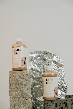 Two bottles labeled 'hella co.' with designs indicating milk tea products, one for shower cream and the other for hair therapy, are placed on textured stone-like blocks. A reflective, crinkled foil serves as the backdrop, adding a metallic element to the composition.