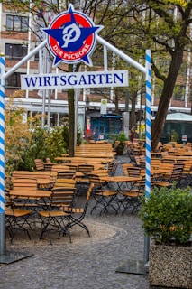 An outdoor beer garden with rows of empty wooden tables and chairs arranged neatly on a cobblestone surface. Above the seating area, a decorative sign reads 'WIRTSGARTEN' with blue and white striped poles on either side. Trees with budding leaves are visible, suggesting a spring setting.