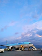 A Korean Air airplane is parked on the tarmac with its cargo hold open. The sky is mostly clear with some scattered clouds, displaying a mix of blue and pastel hues. Ground vehicles and personnel are present around the aircraft.