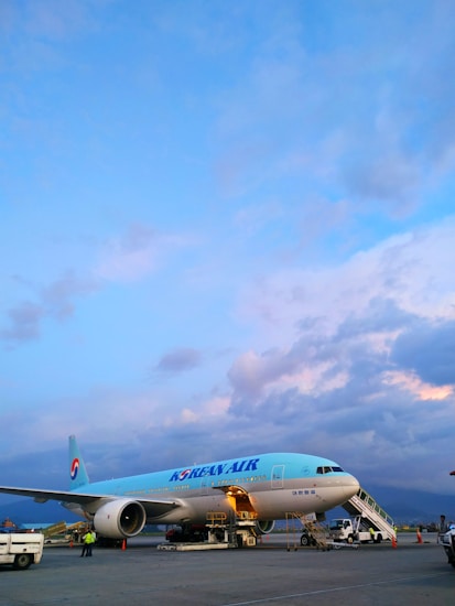 A Korean Air airplane is parked on the tarmac with its cargo hold open. The sky is mostly clear with some scattered clouds, displaying a mix of blue and pastel hues. Ground vehicles and personnel are present around the aircraft.