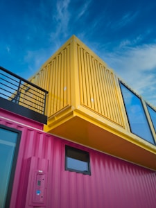 A modern architectural structure made from brightly colored shipping containers is depicted. The building features a pink container at the base with a yellow container stacked on top, both having large glass windows. The vibrant colors contrast with the clear blue sky, giving the structure a contemporary and artistic appearance.