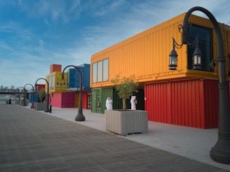A colorful series of shipping containers stacked to create a vibrant urban setting. The foreground features a wide wooden boardwalk, lined with ornamental street lamps. A few trees are planted in large concrete planters, while two people in traditional attire stand near the containers, suggesting a leisurely or casual atmosphere.