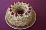 A round cake adorned with creamy swirls and topped with red cherries. It has a light texture, covered with crushed nuts, and sits on a ceramic plate against a maroon backdrop.