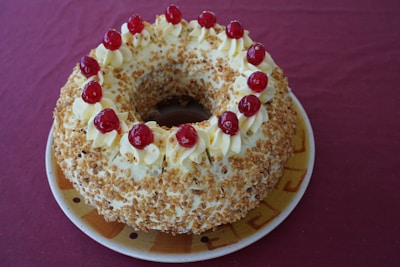 A round cake adorned with creamy swirls and topped with red cherries. It has a light texture, covered with crushed nuts, and sits on a ceramic plate against a maroon backdrop.