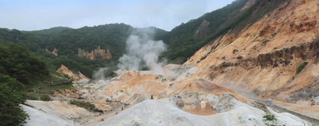 A geothermal landscape with steaming vents and barren, rocky terrain surrounded by lush green hills. The ground is primarily composed of orange, brown, and white hues, indicating sulfur and mineral deposits. Plumes of steam rise from the surface, creating a mystical atmosphere.