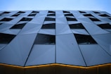 A modern building facade with a geometric, angular pattern of metal panels. The design creates a dynamic visual effect with alternating protruding and recessed shapes. The surface reflects a cool blue tint, possibly due to the lighting or material. The bottom edge features a thin, horizontal line of warm, yellow-orange light, adding contrast to the structure.