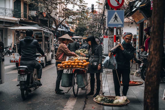 A busy street scene with street vendors selling fruits and vegetables. People are interacting near the stalls, and a man wearing a traditional hat is arranging oranges on a bicycle basket. Several pedestrians and a motorcyclist are passing by. Various signs and storefronts line the bustling urban environment.