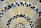 A close-up view of a mosaic pattern consisting of various triangular and quadrilateral pieces. The tiles are arranged in concentric circles, with a mix of creamy beige, dark blue, and black colors. The grout between the tiles is a light color, accentuating the geometric patterns of the mosaic.