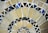 A close-up view of a mosaic pattern consisting of various triangular and quadrilateral pieces. The tiles are arranged in concentric circles, with a mix of creamy beige, dark blue, and black colors. The grout between the tiles is a light color, accentuating the geometric patterns of the mosaic.