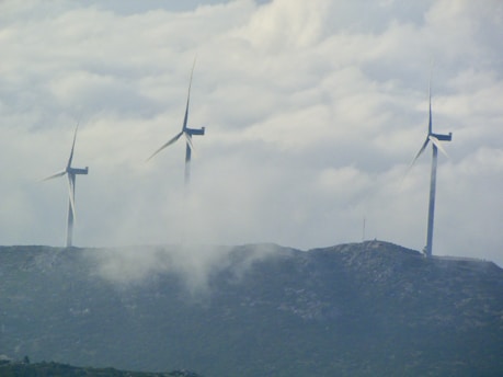 Three large wind turbines stand atop a hilly landscape, partially obscured by drifting clouds. The sky is overcast with dense cloud cover, creating a serene and slightly mysterious atmosphere. The turbines are spaced evenly across the ridge, symbolizing renewable energy and environmental stewardship.
