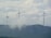 Three large wind turbines stand atop a hilly landscape, partially obscured by drifting clouds. The sky is overcast with dense cloud cover, creating a serene and slightly mysterious atmosphere. The turbines are spaced evenly across the ridge, symbolizing renewable energy and environmental stewardship.