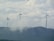 Three large wind turbines stand atop a hilly landscape, partially obscured by drifting clouds. The sky is overcast with dense cloud cover, creating a serene and slightly mysterious atmosphere. The turbines are spaced evenly across the ridge, symbolizing renewable energy and environmental stewardship.