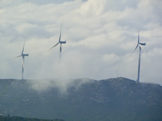 Three large wind turbines stand atop a hilly landscape, partially obscured by drifting clouds. The sky is overcast with dense cloud cover, creating a serene and slightly mysterious atmosphere. The turbines are spaced evenly across the ridge, symbolizing renewable energy and environmental stewardship.