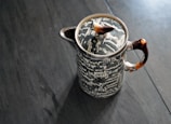 A uniquely patterned ceramic teapot with a black and white geometric design sits on a dark wooden surface. The teapot features a distinct lid handle resembling a small sculpted element and has deep brownish-red handles.