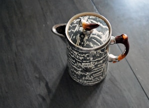 A uniquely patterned ceramic teapot with a black and white geometric design sits on a dark wooden surface. The teapot features a distinct lid handle resembling a small sculpted element and has deep brownish-red handles.