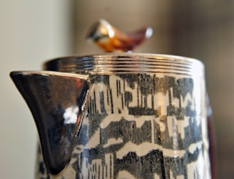 A close-up of a ceramic object featuring a glossy spout and abstract black and white patterns. Perched on the lid is a small, stylized bird figure with a glossy finish.