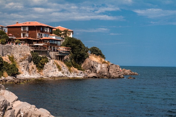 A coastal scene featuring a group of houses perched on rocky cliffs overlooking the ocean. The houses have red tiled roofs and are surrounded by lush greenery. Below the cliffs, the deep blue sea stretches out under a partly cloudy sky.