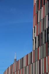 A modern building with a facade featuring vertical panels in shades of red, gray, and beige stretches upward against a clear blue sky. In the distance, a communication tower with a red and white striped antenna emerges. The geometric design of the building creates a dynamic visual rhythm.