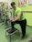 A man wearing a bright green athletic shirt, black pants, and a cap is standing in a gym. He has one foot on a small platform and is smiling at the camera. In the background, there are exercise bikes and artwork on the green walls.