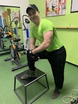 A man wearing a bright green athletic shirt, black pants, and a cap is standing in a gym. He has one foot on a small platform and is smiling at the camera. In the background, there are exercise bikes and artwork on the green walls.
