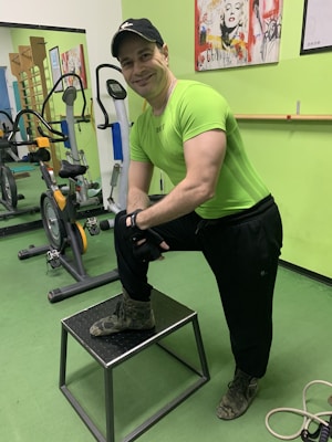 A man wearing a bright green athletic shirt, black pants, and a cap is standing in a gym. He has one foot on a small platform and is smiling at the camera. In the background, there are exercise bikes and artwork on the green walls.
