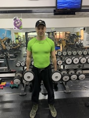A person is standing in a gym holding two dumbbells. The individual is wearing a lime green shirt, black cap, and camouflage boots. The gym is equipped with various weights and machines, visible in the background. There is a cartoon poster on the mirrored wall and a TV screen mounted high above.