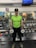 A person is standing in a gym holding two dumbbells. The individual is wearing a lime green shirt, black cap, and camouflage boots. The gym is equipped with various weights and machines, visible in the background. There is a cartoon poster on the mirrored wall and a TV screen mounted high above.