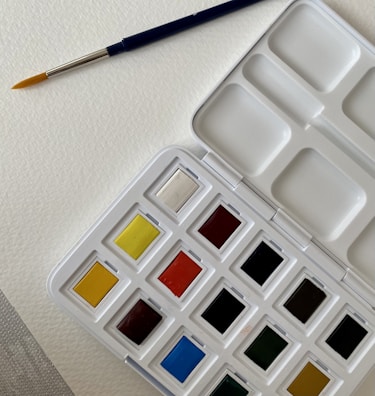 A watercolor paint palette with an assortment of colors in individual pans. A blue-handled paintbrush with a fine tip rests on textured white paper beside the palette.