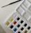 A watercolor paint palette with an assortment of colors in individual pans. A blue-handled paintbrush with a fine tip rests on textured white paper beside the palette.