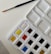 A watercolor paint palette with an assortment of colors in individual pans. A blue-handled paintbrush with a fine tip rests on textured white paper beside the palette.