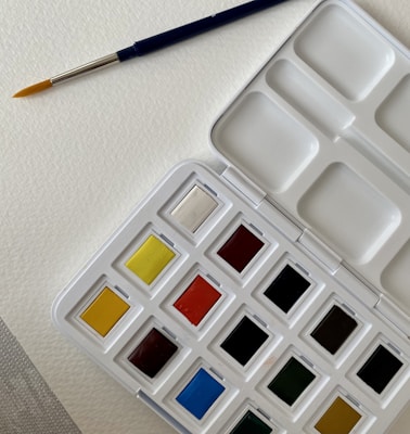 A watercolor paint palette with an assortment of colors in individual pans. A blue-handled paintbrush with a fine tip rests on textured white paper beside the palette.