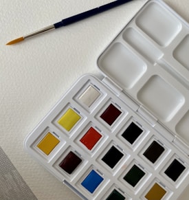 A watercolor paint palette with an assortment of colors in individual pans. A blue-handled paintbrush with a fine tip rests on textured white paper beside the palette.