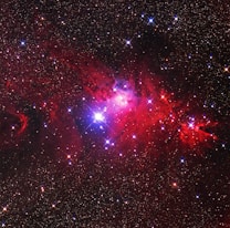 An expansive view of a starry sky features a vibrant nebula. Bright stars punctuate the dark backdrop, shining through clouds of red and purple dust. The nebula is illuminated with intense shades, creating a sense of cosmic wonder.