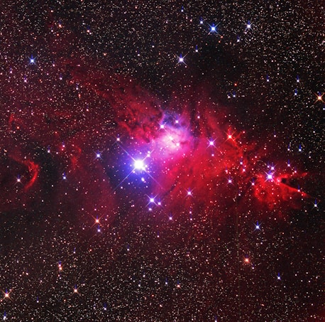 An expansive view of a starry sky features a vibrant nebula. Bright stars punctuate the dark backdrop, shining through clouds of red and purple dust. The nebula is illuminated with intense shades, creating a sense of cosmic wonder.