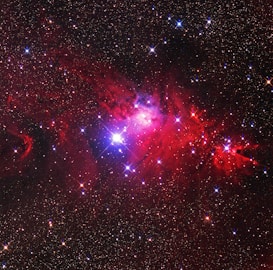 An expansive view of a starry sky features a vibrant nebula. Bright stars punctuate the dark backdrop, shining through clouds of red and purple dust. The nebula is illuminated with intense shades, creating a sense of cosmic wonder.