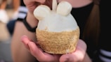 A hand holds a dessert designed to resemble a bear, with a white chocolate coating and light brown crumbly texture resembling fur. The treat is topped with two small round ears.