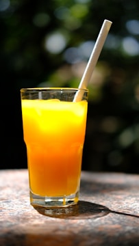 A tall glass filled with bright yellow-orange juice, possibly orange juice, with a white straw. The glass is set on a textured surface with a blurred, greenish background, suggesting an outdoor setting with natural light casting shadows.