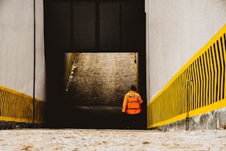 A person wearing an orange high-visibility jacket with a logo on the back walks away from the camera down a narrow passageway. The passage is bordered by tall concrete walls with yellow and black striped warning patterns.