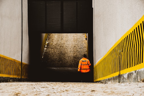 A person wearing an orange high-visibility jacket with a logo on the back walks away from the camera down a narrow passageway. The passage is bordered by tall concrete walls with yellow and black striped warning patterns.