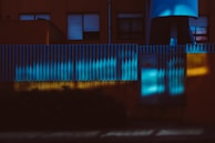 A building facade with a modern, industrial design features vertical, ribbed metal paneling illuminated by blue and yellow light. The lights cast intriguing patterns and shadows across the surfaces, creating a dramatic visual effect. Windows with partially closed blinds are visible, adding to the geometric aesthetic.