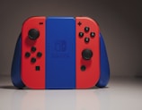 A Nintendo Switch console is set against a neutral gray background. The console features the iconic red and blue Joy-Con controllers on either side of the central blue unit, with the Nintendo Switch logo visible.