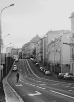 A black and white photograph featuring an urban street scene. The street stretches uphill, lined with parked cars on either side. Architectural buildings with classic designs are visible, along with a lone cyclist traveling towards the camera. The sky is overcast, contributing to the subdued atmosphere.