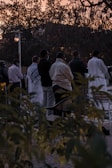 A group of people standing in an outdoor setting during sunset. They appear to be engaged in a communal or social activity. The background shows trees silhouetted against the evening sky, and the ground is partially obscured by bushes in the foreground.